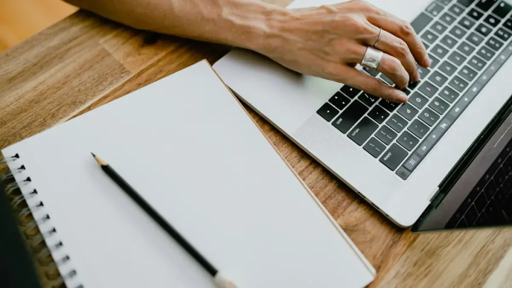 Mains sur le clavier d'un ordinateur portable, à côté d'un carnet ouvert et d'un crayon