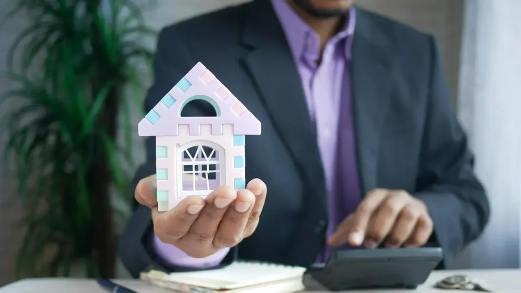 Homme en costume tenant une maquette de maison au-dessus d'une calculatrice