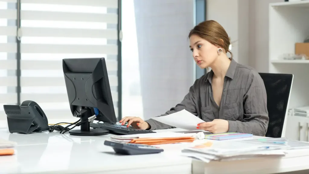 Femme qui travaille à son bureau devant un écran, des documents à la main